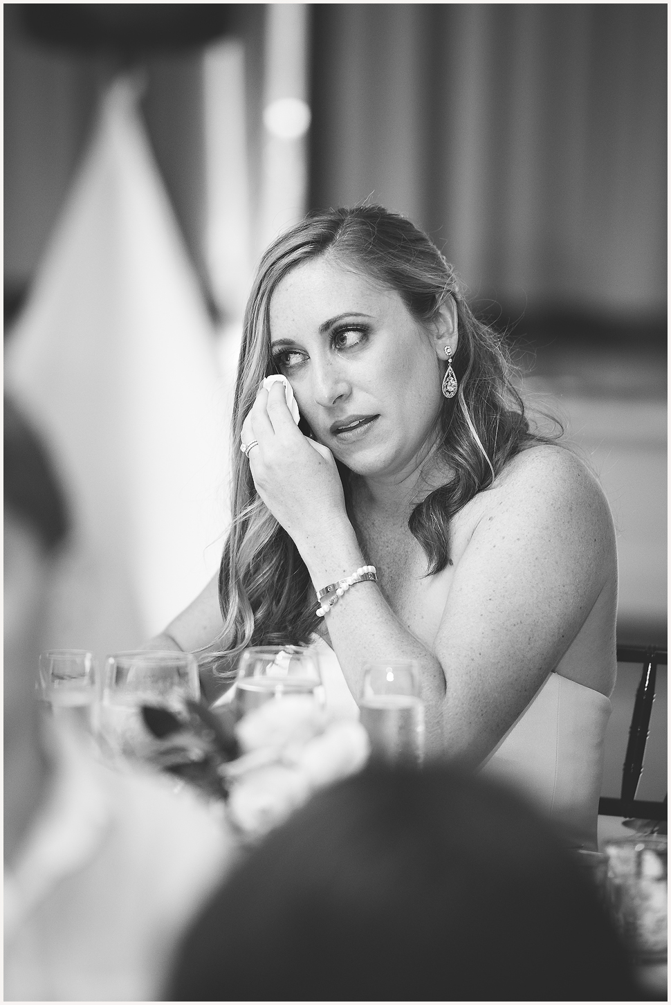 Black and white candid photo of a bride wiping a tear from her face while listening to the speeches at her wedding. 