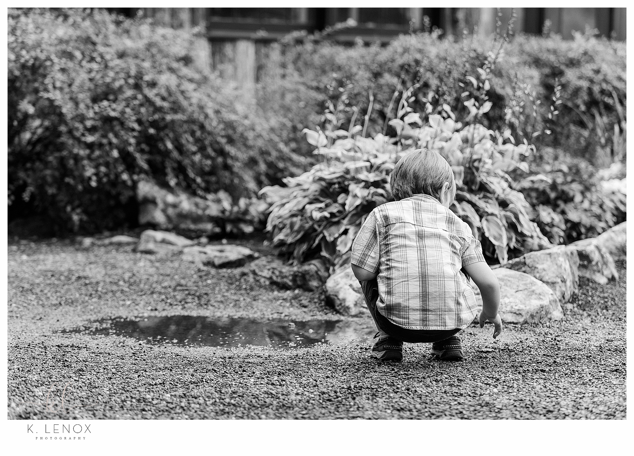 Black and White candid photo of a young boy playing in a mud puddle. 