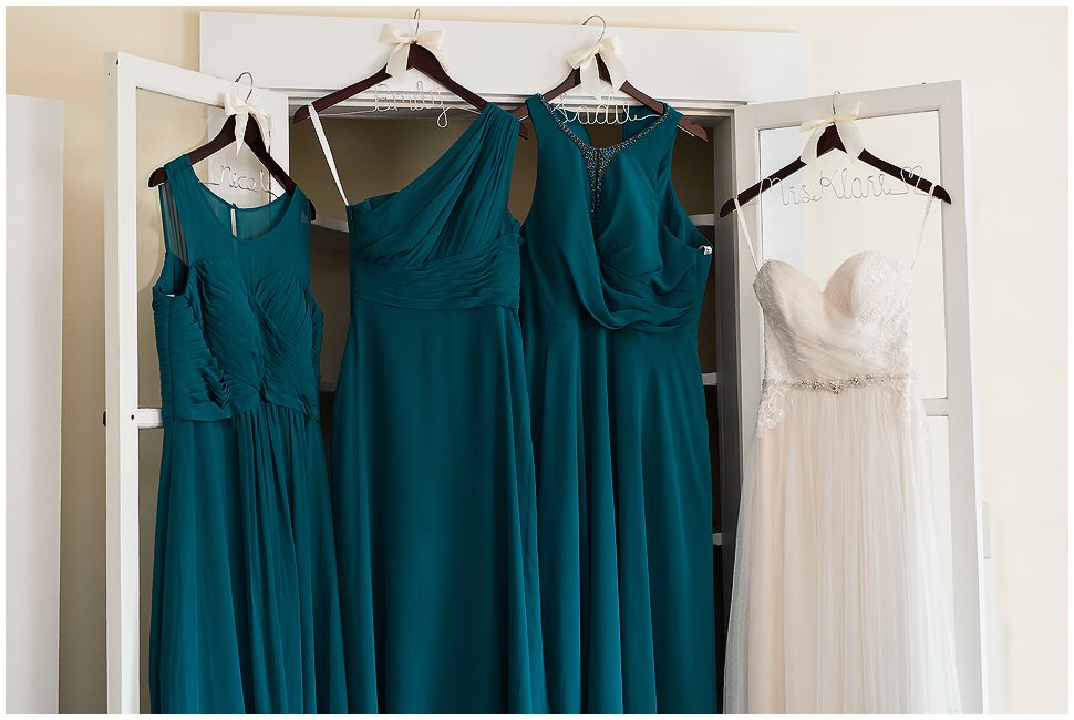 Deep teal Bridesmaids dressings hanging next to simple white wedding dress at Alyson's Orchard. 