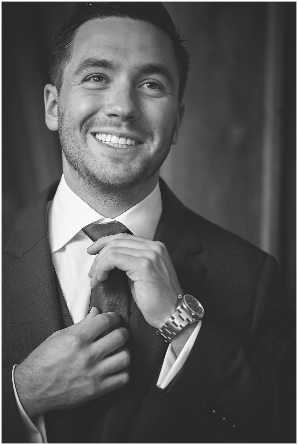 Black and white photo of a groom with scruff fixing his tie. 