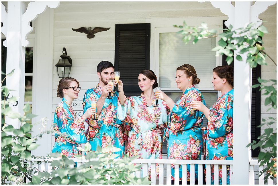 Bridesmaids wearing blue floral robes toast with Mimosa's on the Porch of Alyson's orchard farm house. 