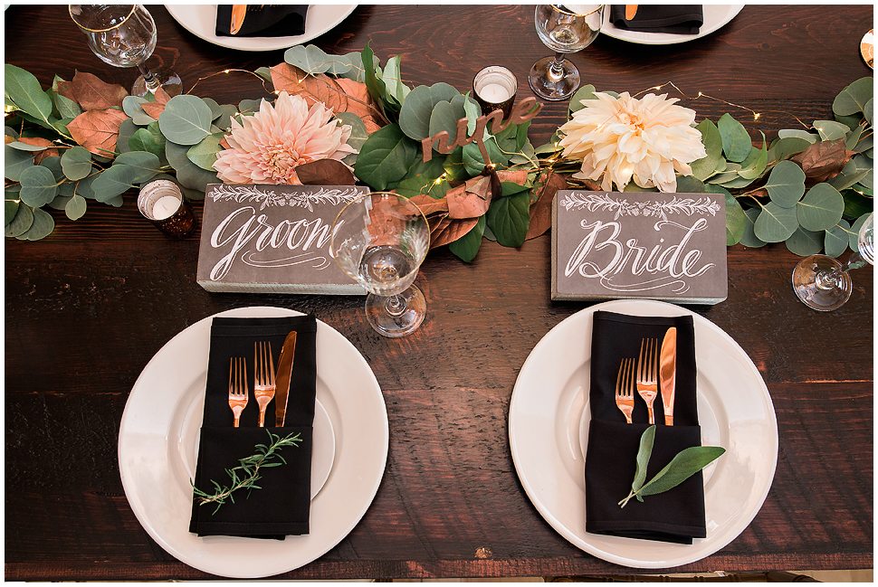 Table scape of a bride and grooms seats showing copper flatware, black napkins, eucalyptus garland. 