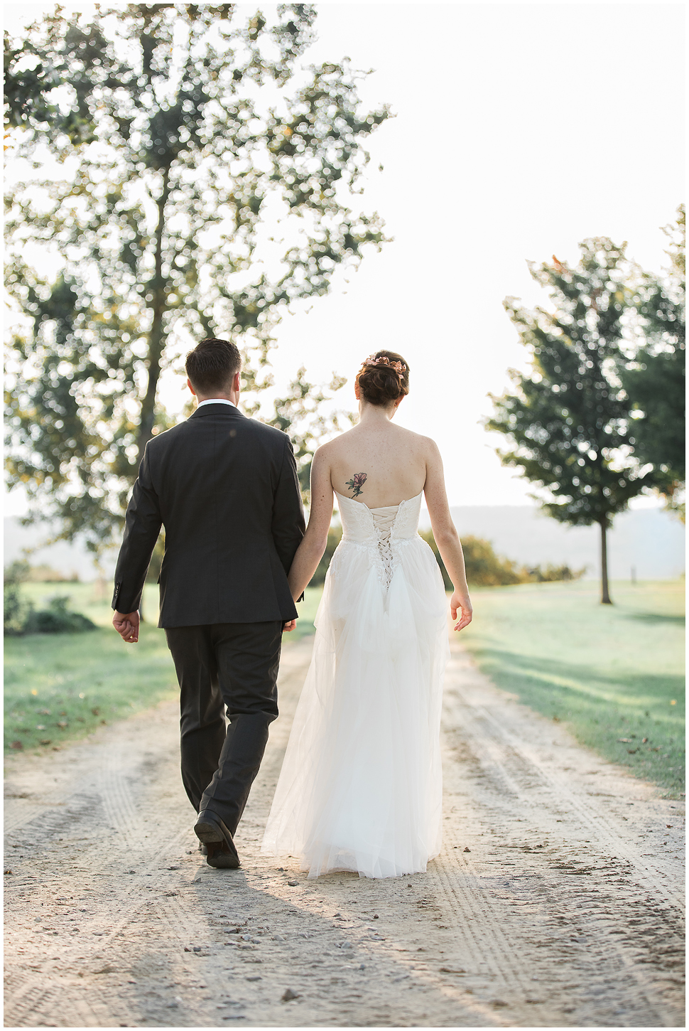 Bride and groom walk hand in hand on a dirt path. Shown from behind. 