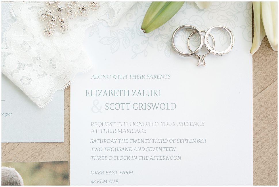Photo shows a close up view of a simple white wedding invitation with light gray writing. Also three wedding rings. 