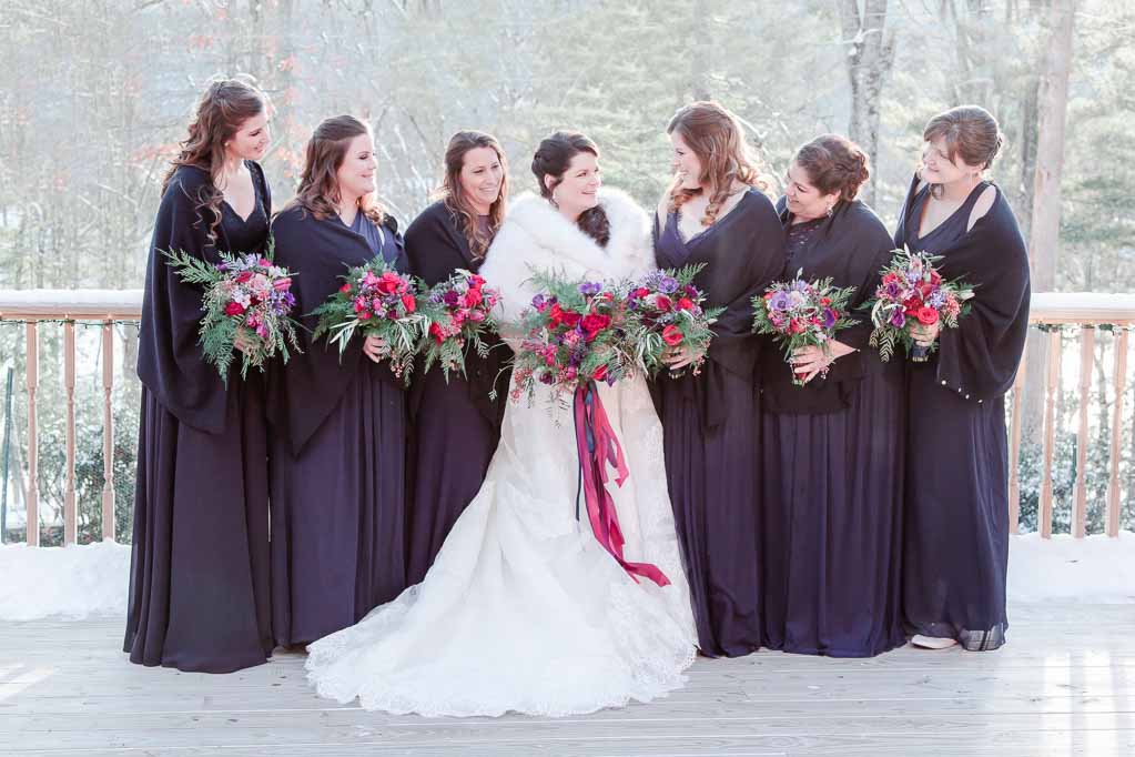 Photo of Bride and 6 bridesmaids wearing deep purple dresses holding wedding bouquets with purple and pink flowers. 