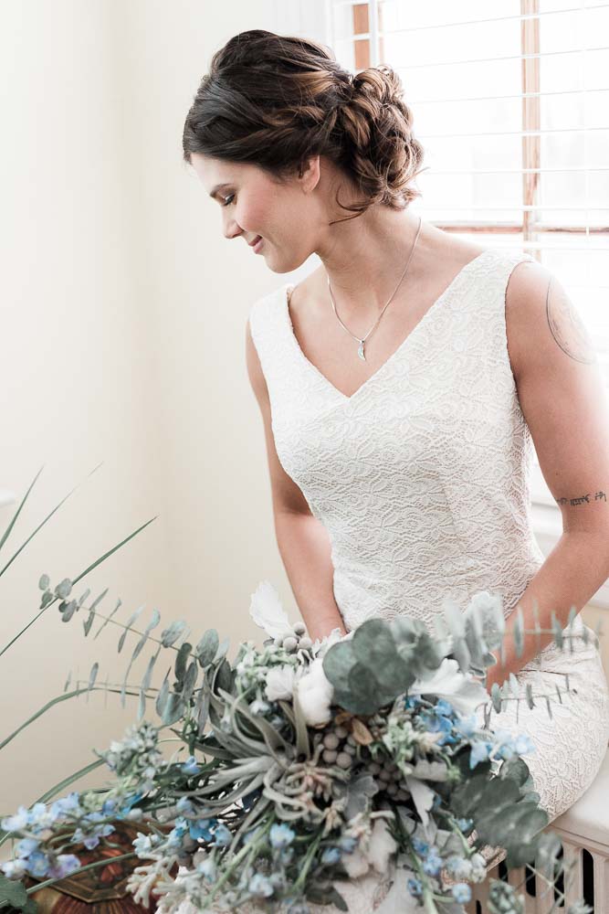 Bride wearing a simple white embroidered dress and holding a bouquet of blue and white flowers with lots of greenery. 