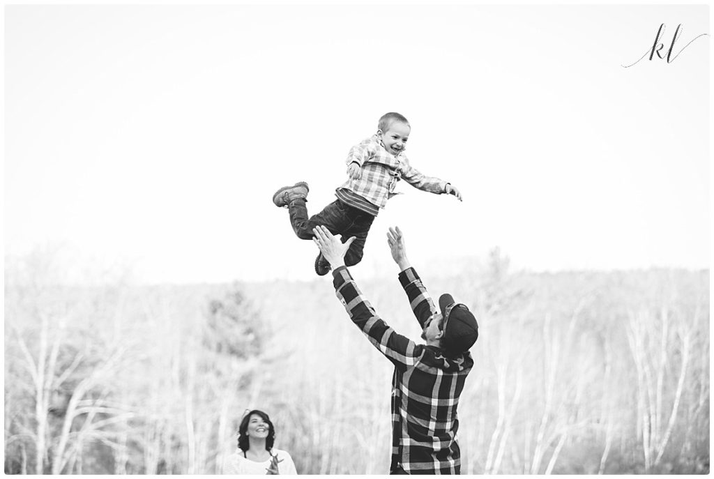 Black and White photo of An authentic moment with a dad throwing his son in the air with mom happily watching. 