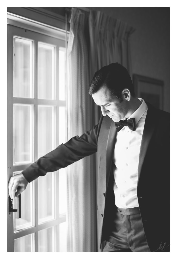Black and White photo of a groom wearing a tuxedo standing in front of his room window at his Bedford Village Inn summer wedding. 