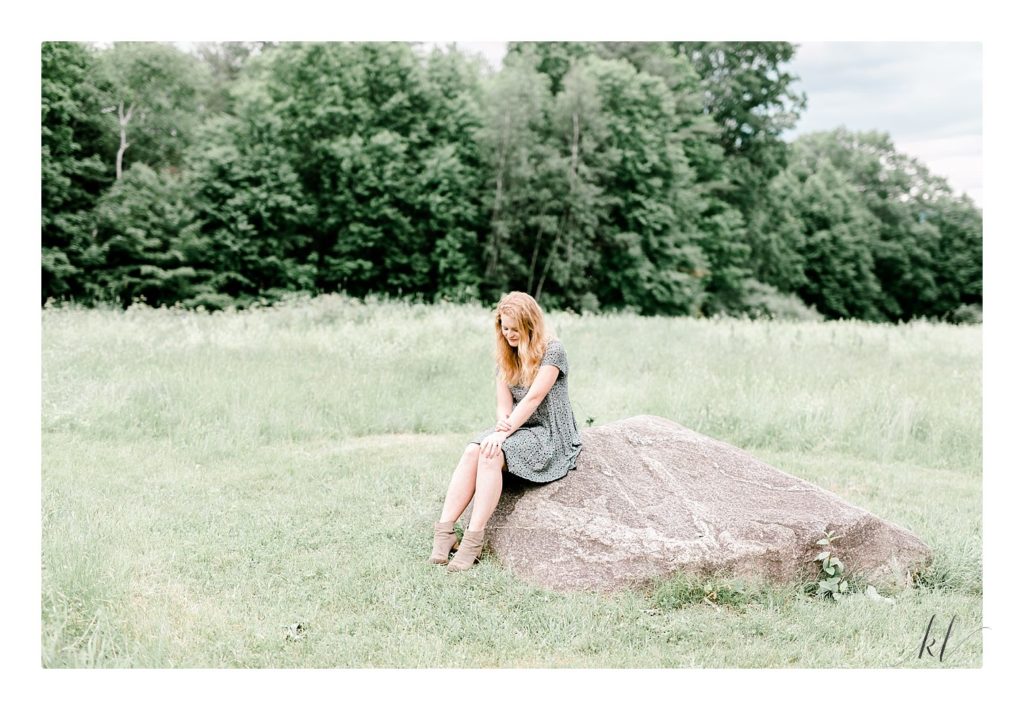 Light and Airy Keene Photographer K. Lenox showcases a blonde senior girl's portrait sitting on a rock in a field. 
