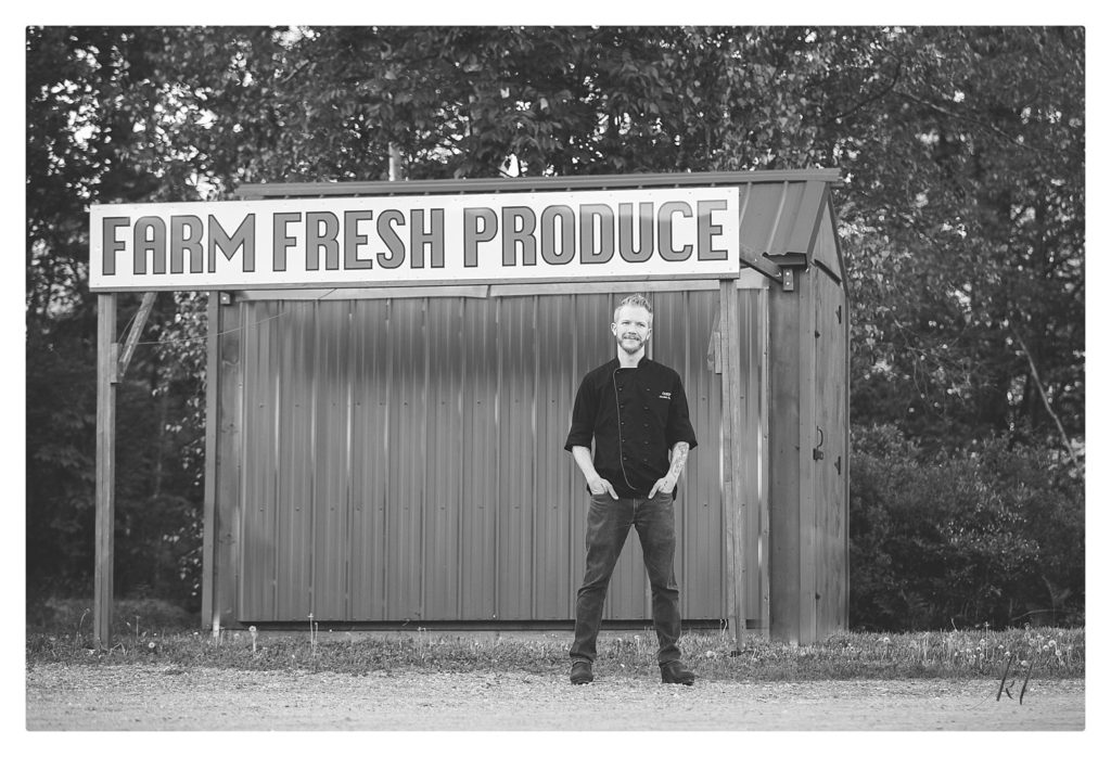 Black and White image of Chef Jordan Scott at Borealis Farm in front of a Farm Fresh Produce sign. 