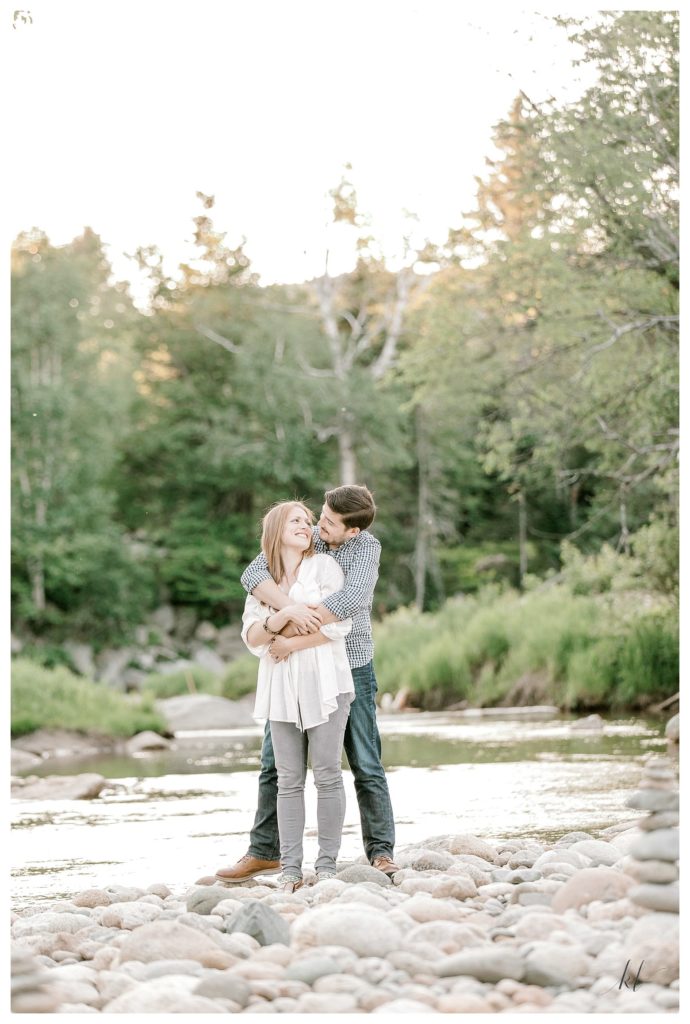Man hugs his fiance from behind on the side of a creek in the White Mountains region of NH. Taken by K. Lenox Photography. 