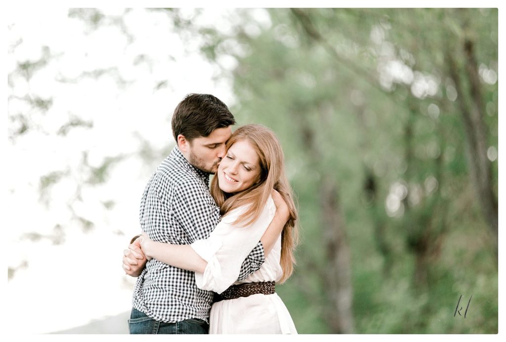 Light and Airy engagement photo of a Man and woman hugging while the man kisses the cheek. 