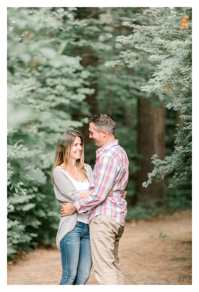 Rustic Engagement Photo of a man and woman smiling at each on a path in the woods. 