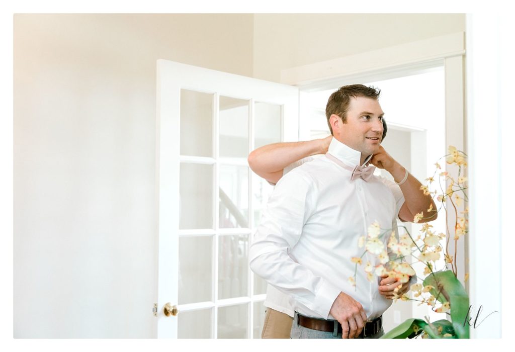 Light and Airy photo of a groom putting on his pink bow tie with help of his friend. 