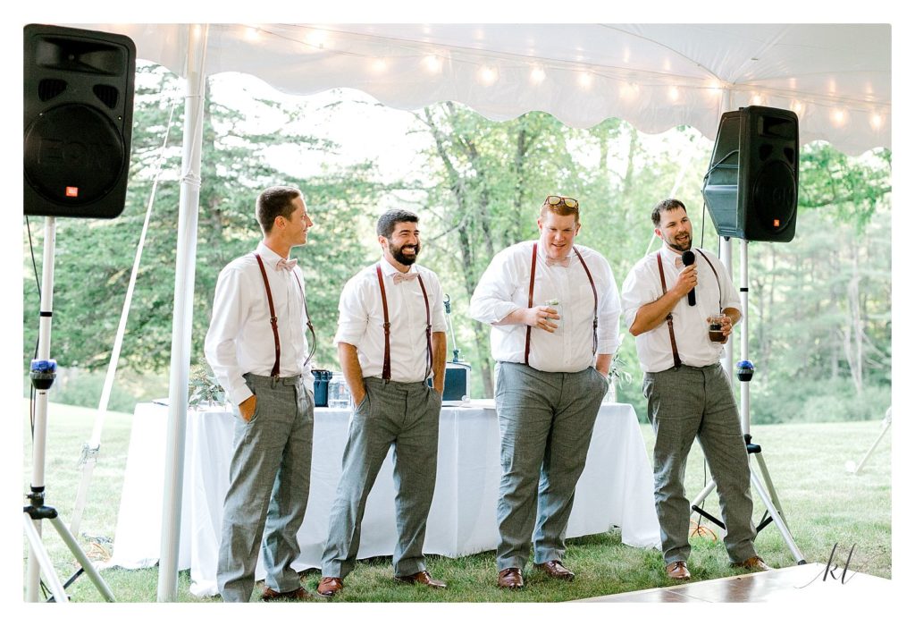 Four Groomsmen wearing leather suspenders, white shirts and gray pants give a speech during a casually elegant wedding reception. 