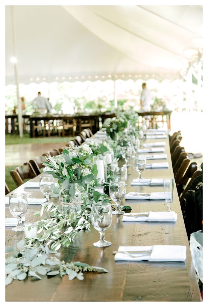 Rustic Farm table wedding table scape showing lots of greenery and small white roses, white candles. 
