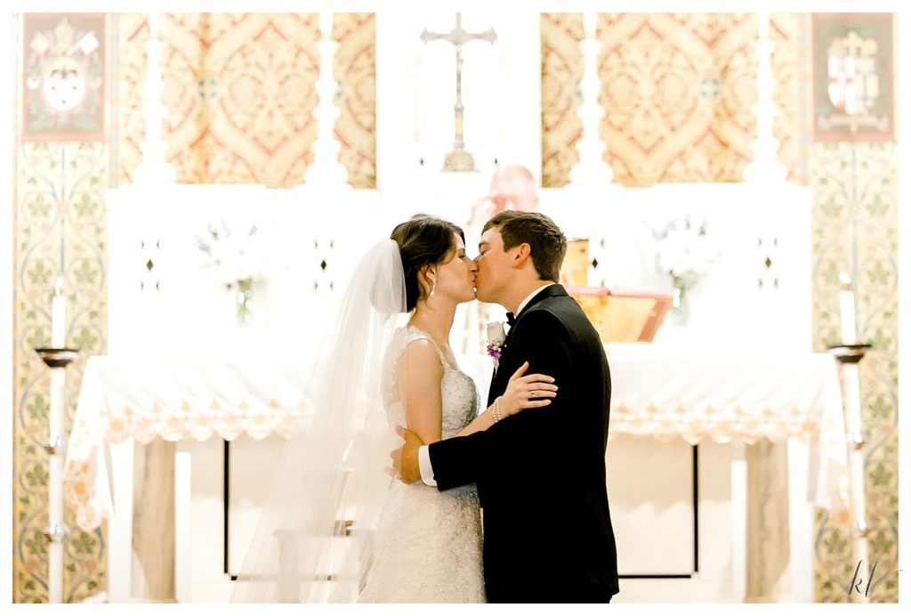 Bride and groom share a first kiss during their Catholic wedding Ceremony in a dark church. 
