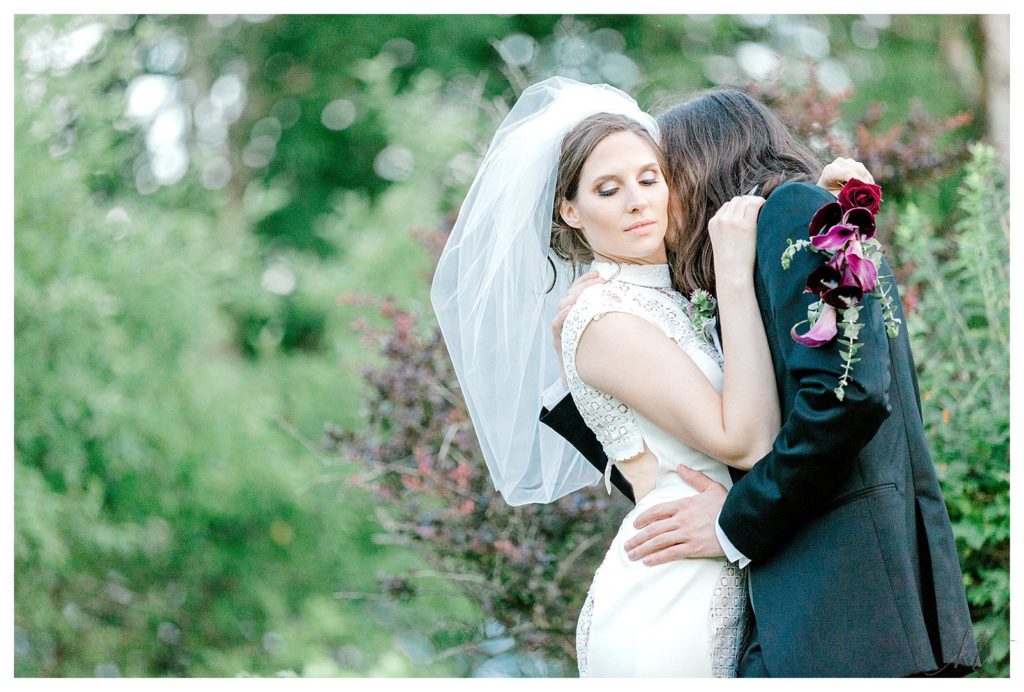 Bride wearing bubble veil and modern lace wedding dress holds Calla Lilly bouquet and hugs her husband. 