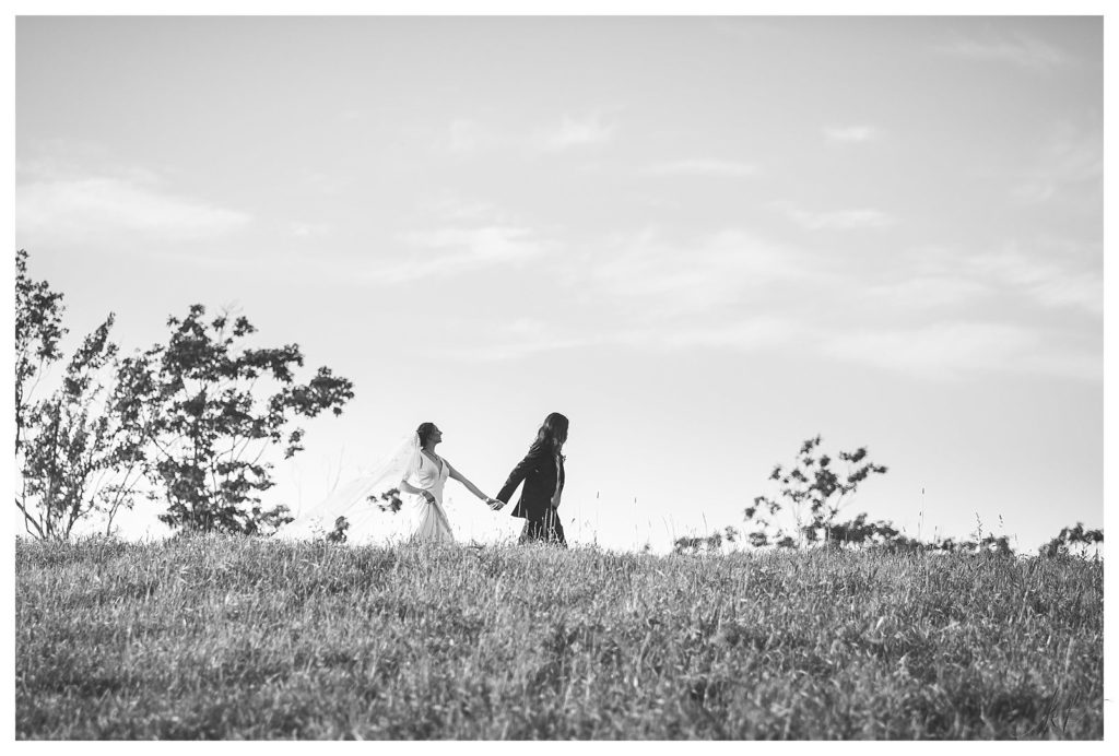 Black and white candid photo of a bride and groom holding hands walking on a hill at mayfair farm. 