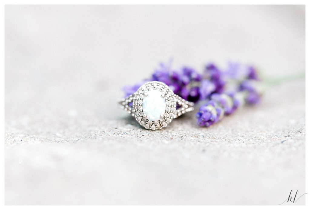 Ring with a soft blue center surrounded by small diamonds. Photographed with purple flowers. 