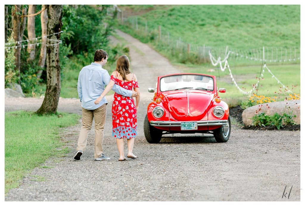 Engaged couple walk with their arms around each other towards their 1972 Red Volks Wagon Beetle. 