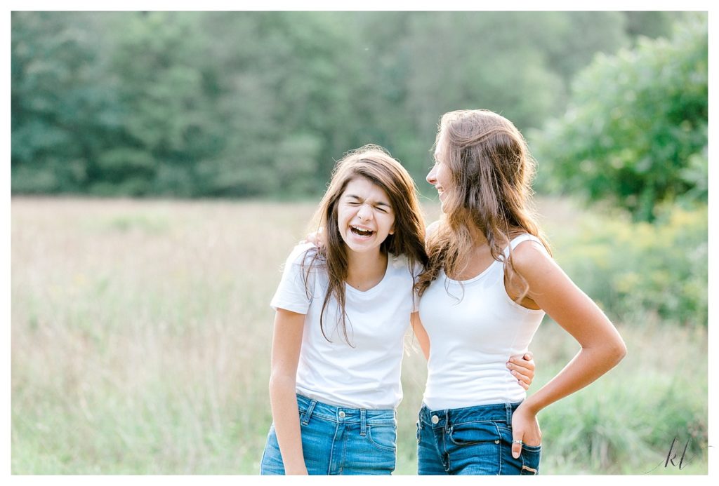 Candid, fun and silly photo of two girls laughing while wearing jeans and white tank-top, standing in a field. 