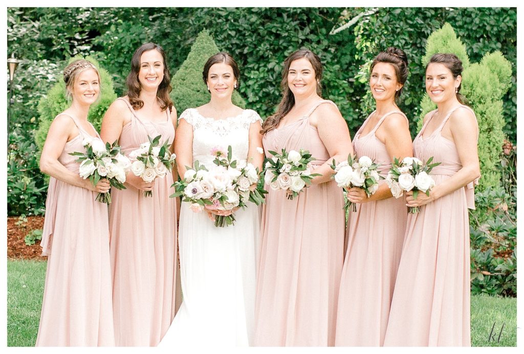Bridesmaids wearing light pink dresses stands with the bride wearing a Stella York Design. Holding floral bouquets. 