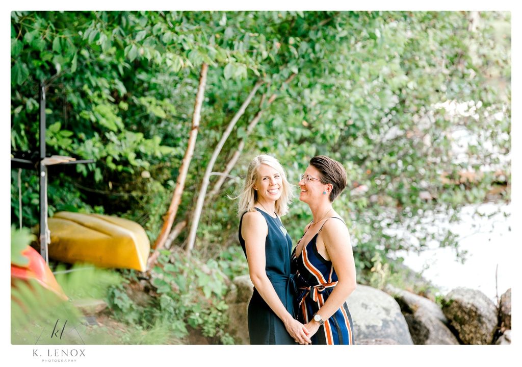 Same Sex couple- engaged women holding hands and smiling on the shore of Lake Sunapee in NH. 