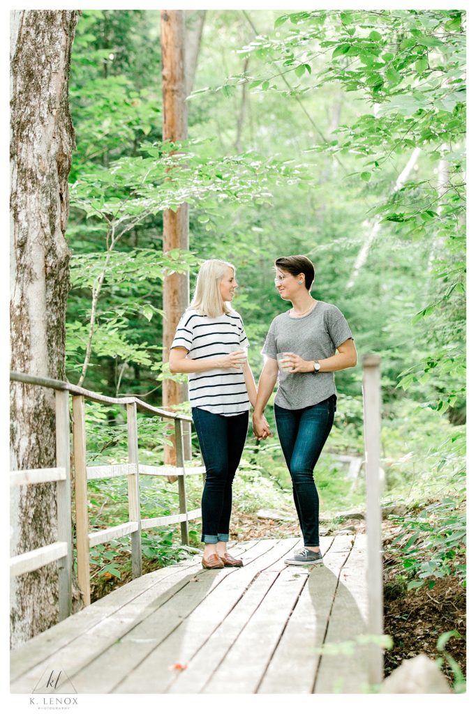 Same sex engaged women holding hands on a wooden foot bridge in the woods near Lake Sunapee NH. Candid engagement photo. 