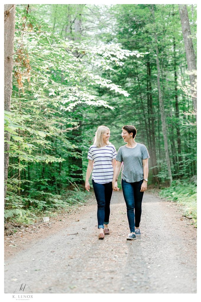 Candid Light and Airy engagement photo using OCF. Two women walk hand in hand on a dirt road surrounded by trees. 