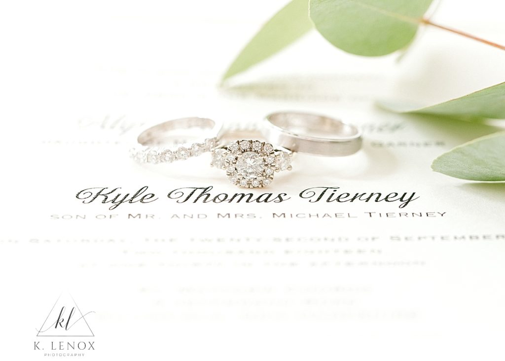 White Invitation with Black cursive lettering with a White Gold diamond Engagement ring and two wedding bands. 