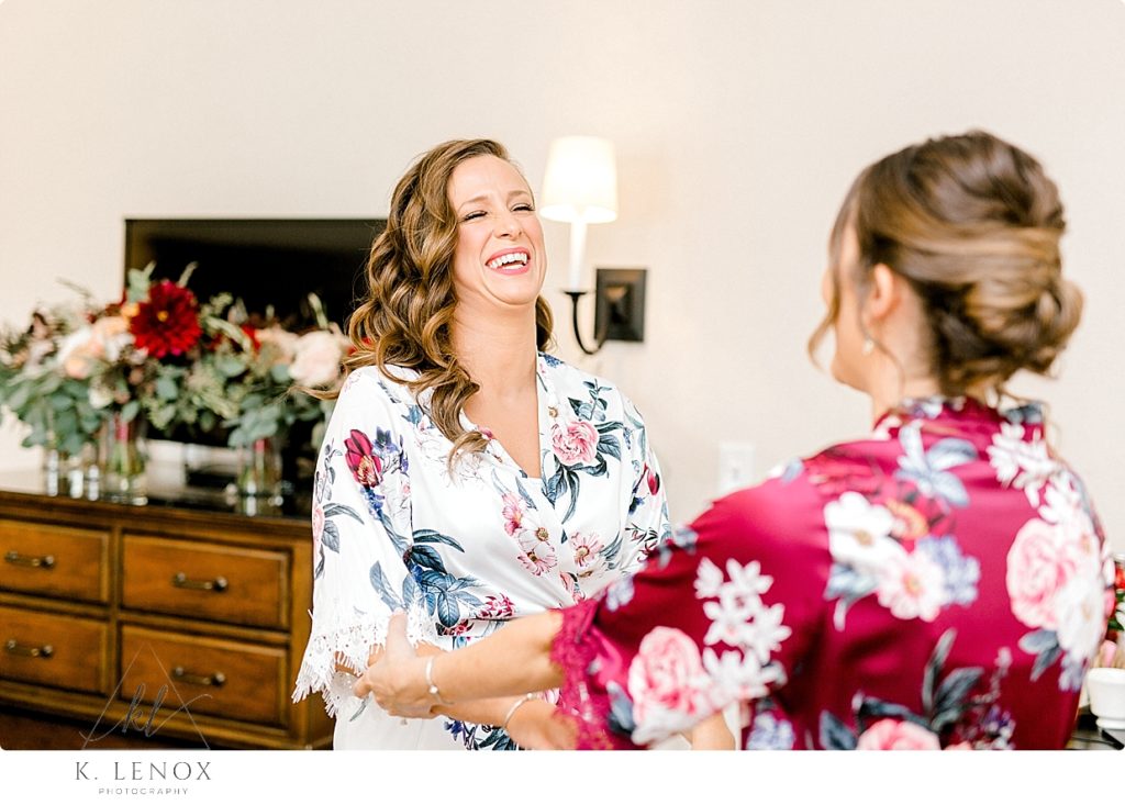 Bride wearing a white floral robe laughs with her bridesmaid. 