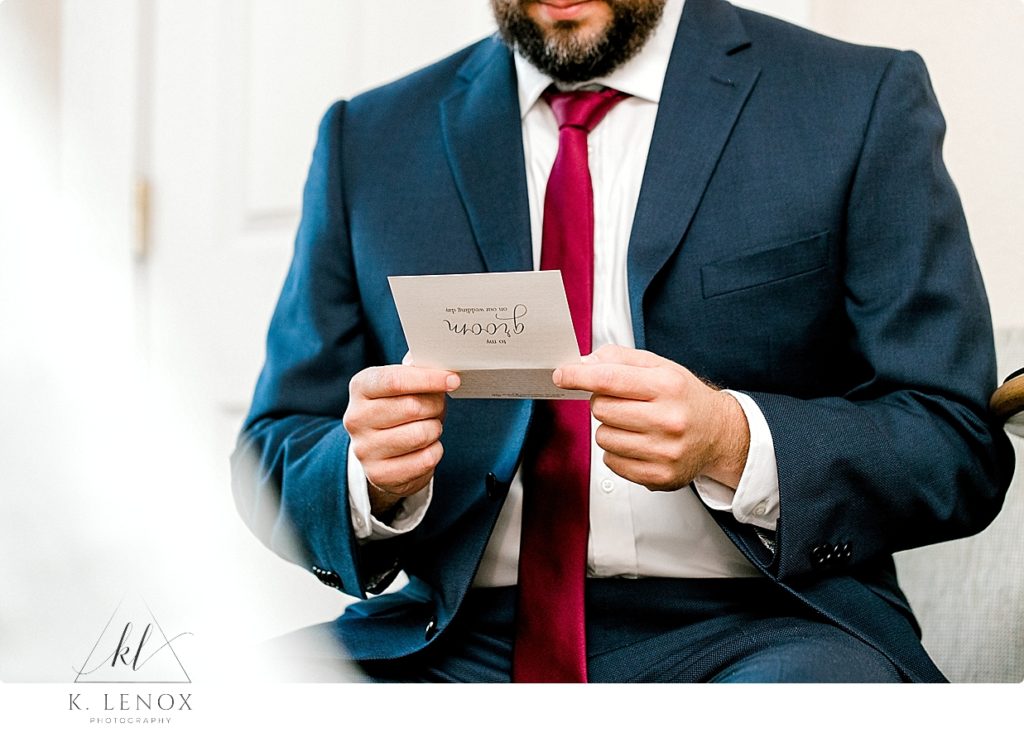 Groom wearing a deep navy blue suit with a maroon tie reads a letter from his bride. 