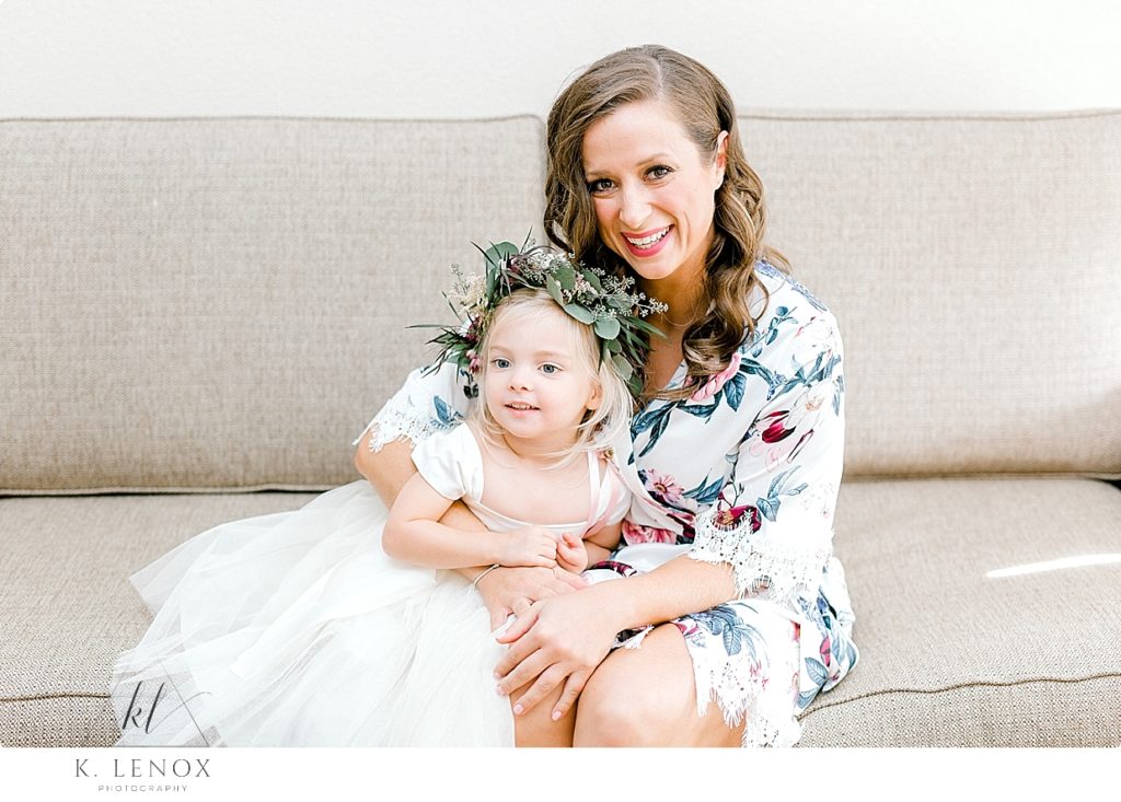 Bride wearing a white floral robe poses for a portrait with her flower girl wearing a floral crown. 