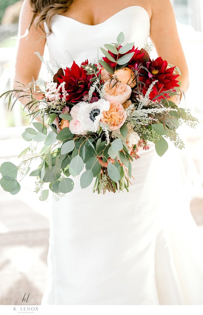 Detail photo showing a bride holding a full bouquet with Fall inspired red flowers, peach flowers white flowers and greenery. 