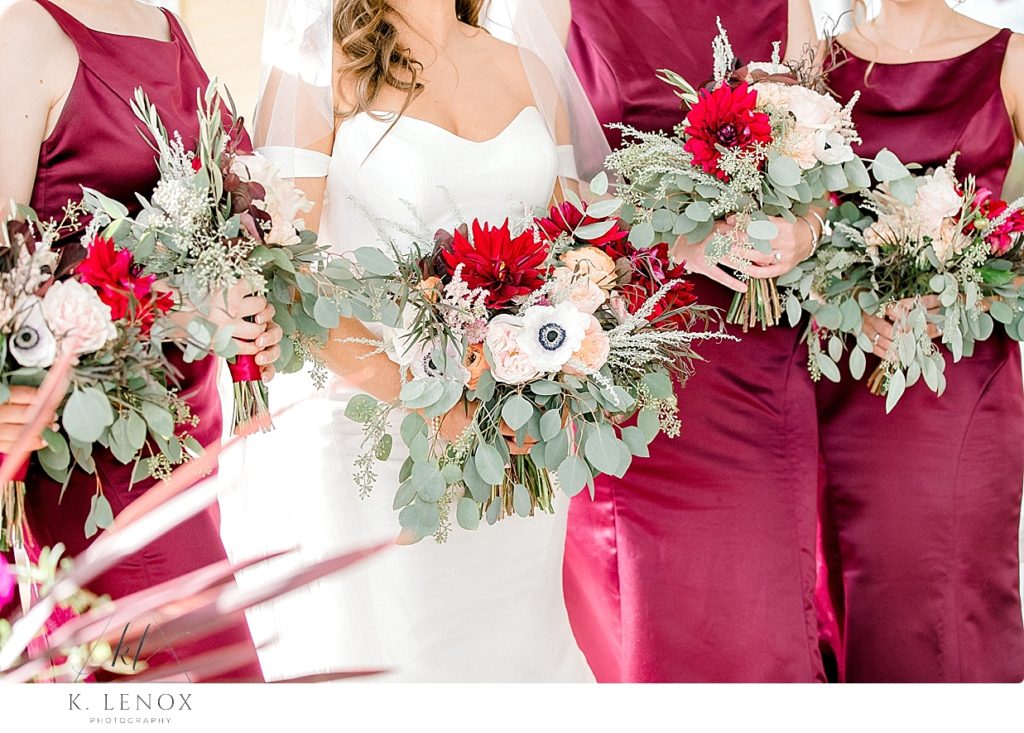 Bride and bridesmaids in Maroon dresses holding floral bouquets with red, white and peach flowers and greenery. 