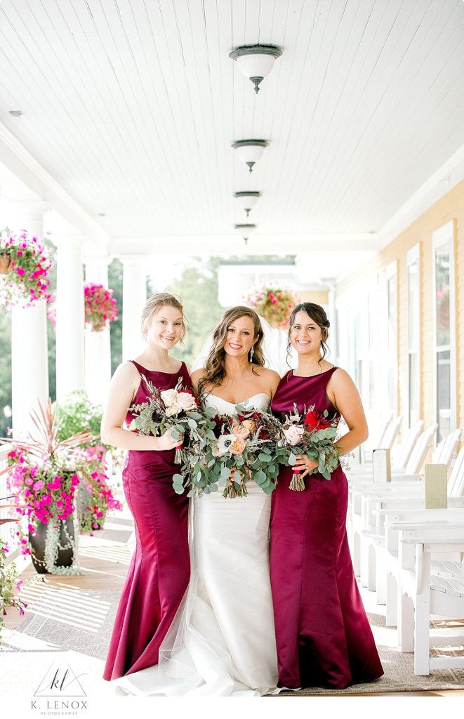 A bride with two bridesmaids wearing maroon gowns stand on the porch of the Mountain View Grand. 