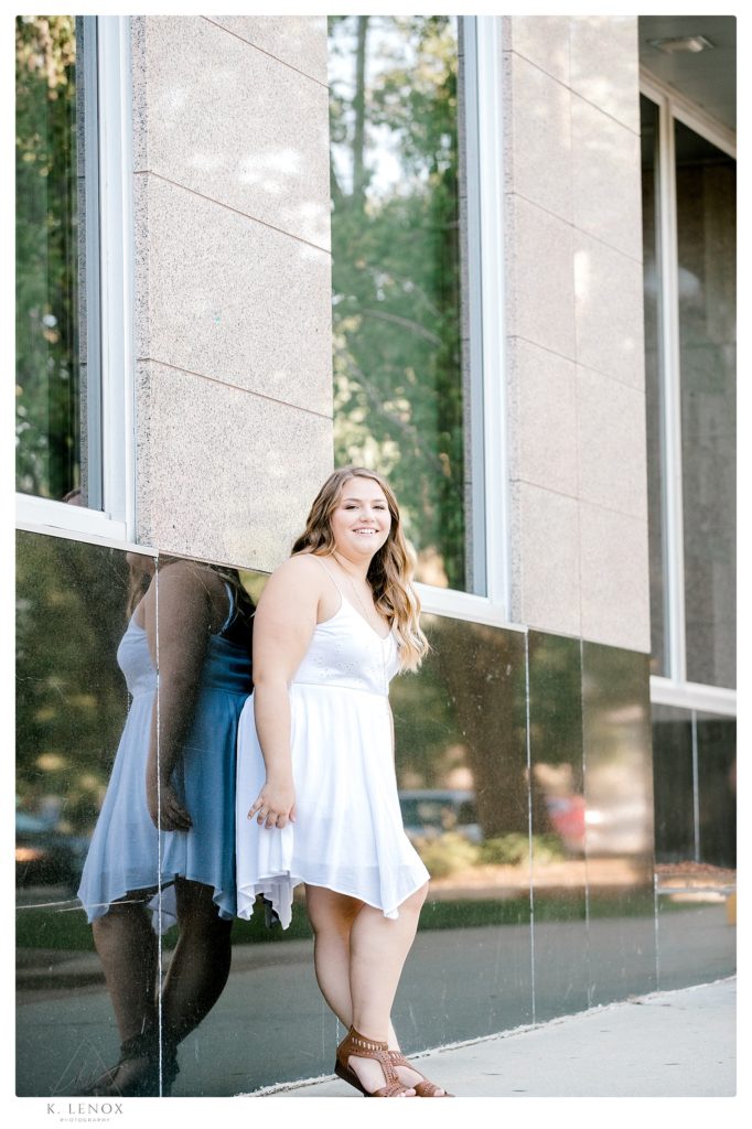 Senior photo in Keene-  Girl wearing a simple white summer dress leans on a wall.  