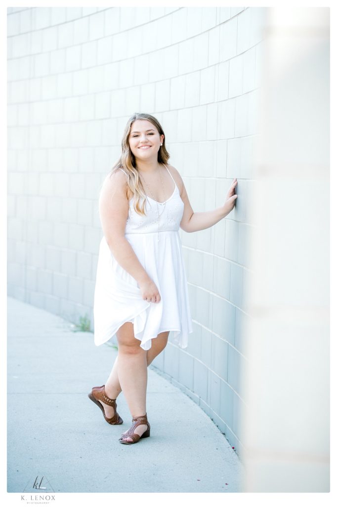 High school senior photo of a girl wearing a white summer dress standing next to the gray wall in Keene NH.   Senior photos in Keene
