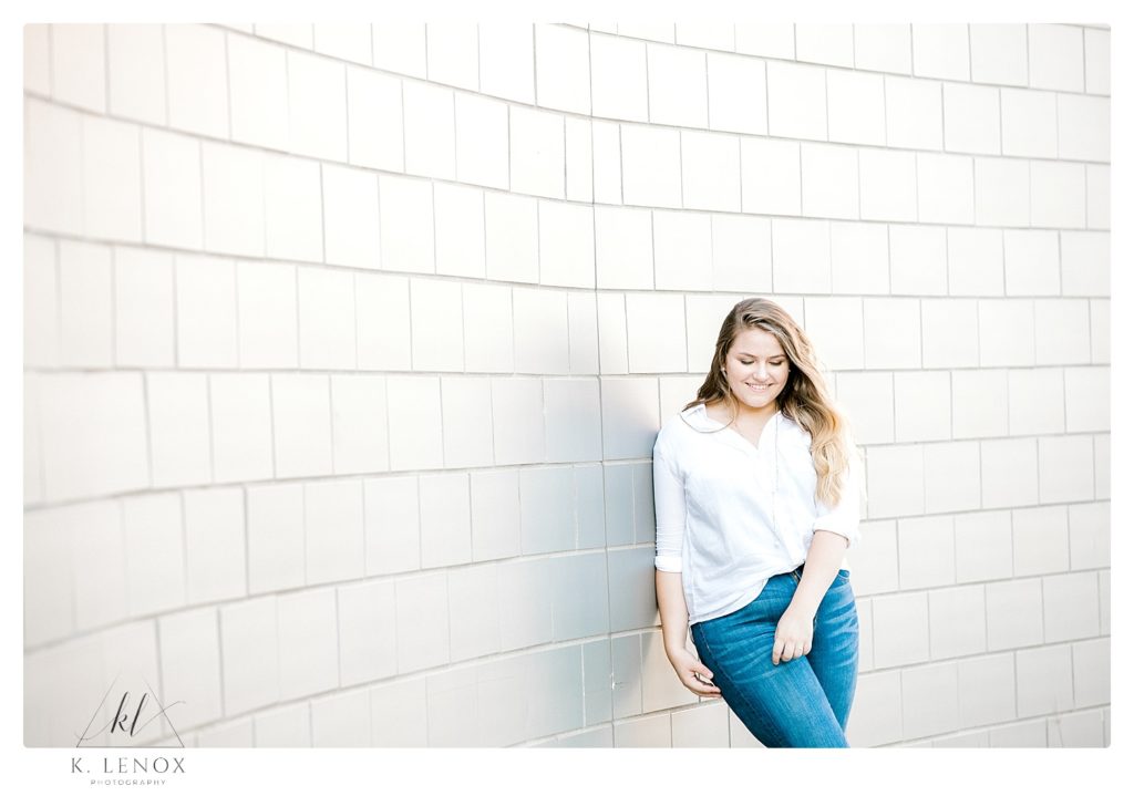 Candid, light and airy senior photo of a girl leaning against a block wall while wearing a white button shirt and blue jeans. 