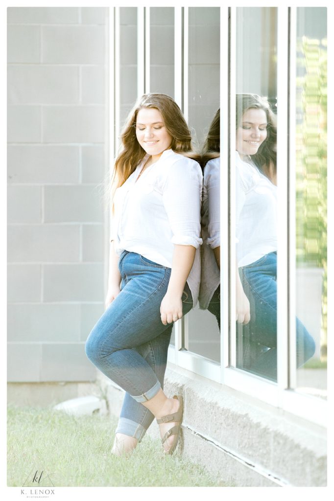 Light and airy senior photo of a girl wearing blue jeans, a white button down shirt standing next to a reflective window. 