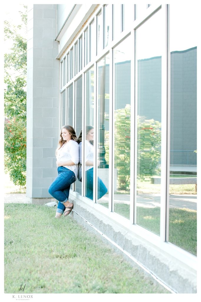 Senior portrait of a girl wearing blue jeans and a white shirt leaning up against a panel of reflective windows. 