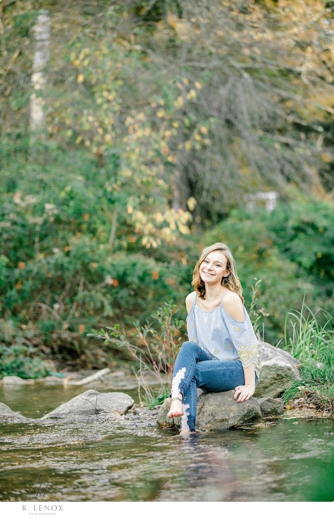 Light and Airy Senior Photo of a girl wearing blue jeans- sitting on a rock near the bank of a stream at Stonewall Farm. 