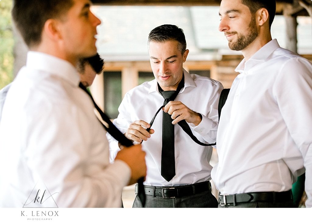 A groom helps his groomsmen tie their black ties, while on the porch of a rustic cabin on Swanzey Lake. 