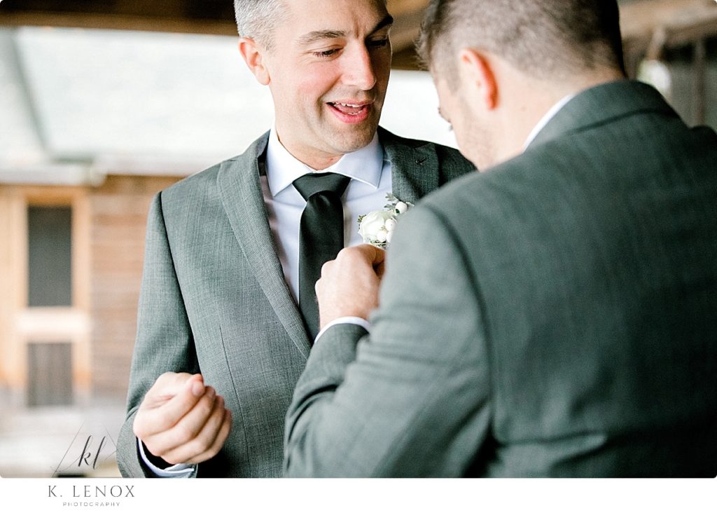 A Man helps a groom button on his Boutonniere on the lapel of his gray suit jacket. 