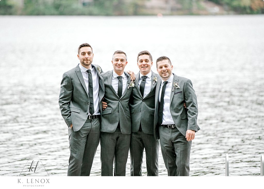 A groom and his three bothers wearing grey suits pose for an informal photo in front of a lake. 