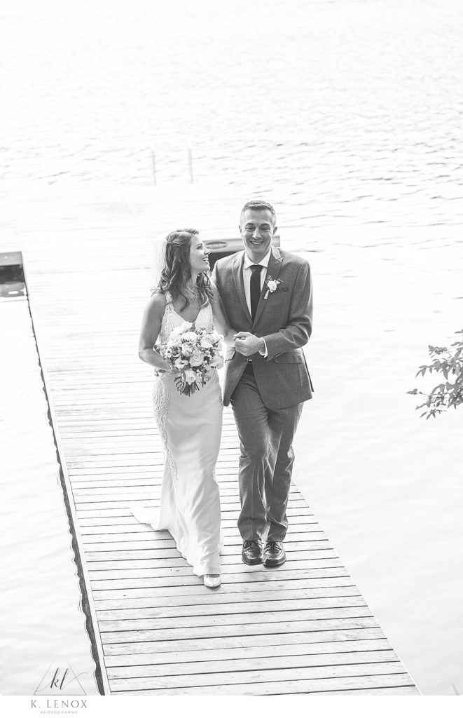 Black and White photo of a groom helping his bride walk on a dock at their lakeside home in Swanzey NH. 