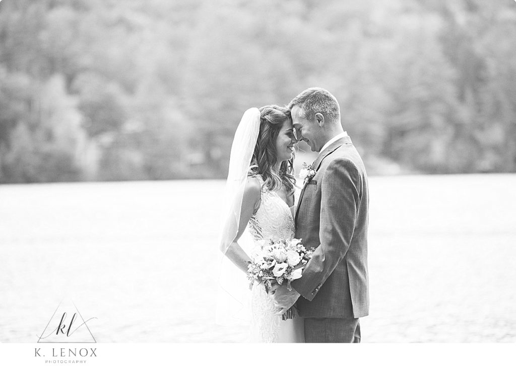 Black and white photo of a bride and groom with forehead to forehead next to the lake. 
