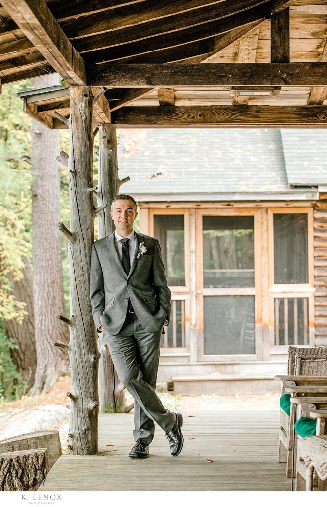 A groom wearing a grey suit poses for a portrait on a rustic lakeside cabin porch. 