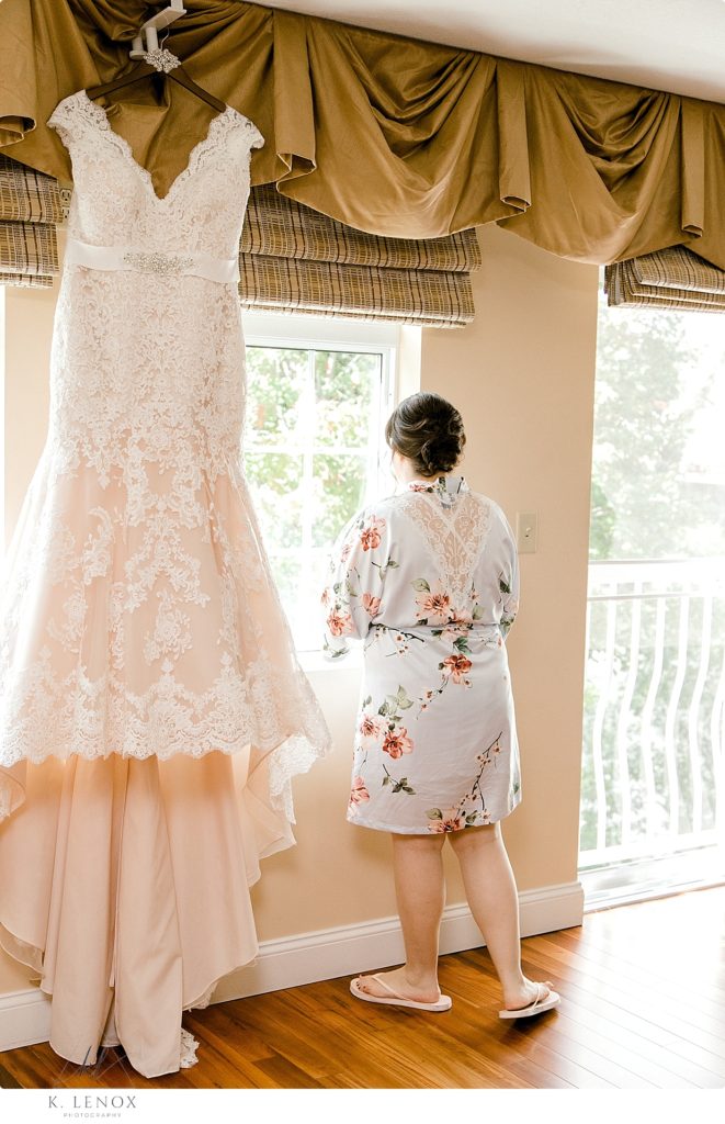 Bride stands next to her wedding dress as she looks out the window at the Riverside Hotel in chesterfield NH. 